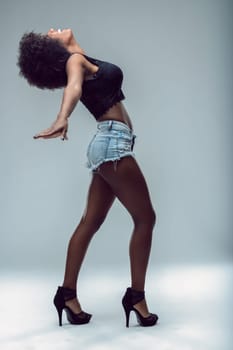 Young latin american woman with afro hairstyle, wearing denim shorts and high heels, is dancing with arms outstretched and head tilted back, expressing joy and freedom