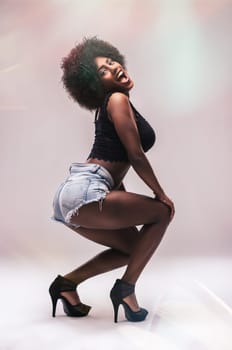 Young black woman with afro hairstyle wearing denim shorts and high heels is dancing and having fun in a studio, looking at the camera with a playful expression