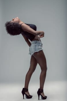 Young latin american woman with curly hair, wearing denim shorts and a black top, is posing in a studio, bending her back and looking up, expressing confidence and sensuality