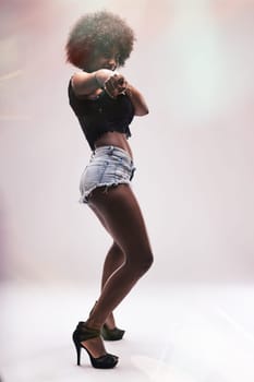 Young woman dancer with vibrant afro hair, wearing stylish denim shorts and high heels, smiling brightly while pointing directly at the camera, exuding confidence and joy