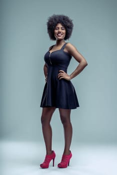 Studio shot of a cheerful young latin american woman with afro hairstyle wearing a black dress and red high heels, posing with hands on her hips and smiling at the camera