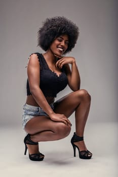 Studio shot featuring a beautiful young black woman with an afro hairstyle, crouching and smiling at the camera while wearing stylish denim shorts and a sleek black top