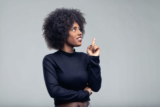 Young woman with vibrant afro hair looking upward and pointing, expressing a moment of inspiration and creativity, isolated against a smooth gray background