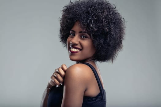 Studio portrait showcasing a beautiful young black woman with a curly afro hairstyle, smiling joyfully over her shoulder against a soft gray background, radiating confidence and happiness