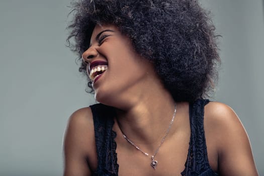 Studio portrait of a beautiful young latin american woman with big frizzy hair, wearing a black top, laughing with a big smile, showing her teeth, on a gray background