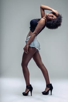 Young woman with an afro hairstyle, wearing stylish denim shorts and high heels, posing confidently while smiling and playfully touching her hair against a gray background