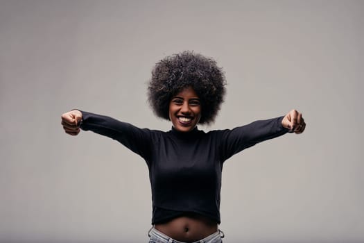 Cheerful young african american woman with big curly hair, wearing a black turtleneck, smiling and stretching her arms forward with clenched fists, radiating strength and happiness