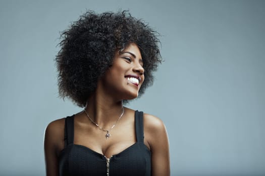 Studio portrait of a cheerful young latin american woman with curly hair, wearing a black top and a necklace, smiling and looking away against a gray background