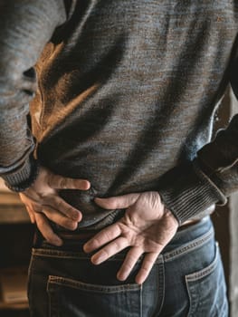 A person discreetly holds their back in pain, dressed casually in a textured sweater and trousers, highlighting everyday discomfort