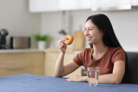 Asian woman eating doughnut contemplating in the kitchen at home