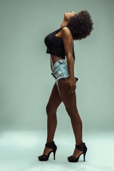 Young latin american model with afro hairstyle posing in a studio, wearing denim shorts, black top and high heels, expressing freedom and confidence