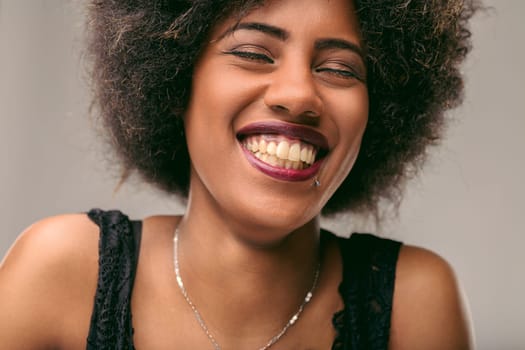Close-up portrait of a cheerful young latin american woman with curly hair, biting her lip and smiling with closed eyes, expressing freedom and self-confidence