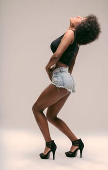 Young latin american woman with afro hair, wearing denim shorts and high heels, striking a confident pose, embodying freedom and diva energy in a studio setting