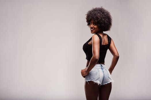 Studio shot of a beautiful smiling latin woman with afro hair, wearing denim shorts and black top, looking back over her shoulder with hands on hips, feeling confident and free