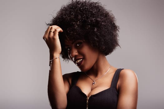 Studio portrait featuring a beautiful young latin american woman smiling while touching her curly afro hair, exuding confidence and sensuality against a soft gray background