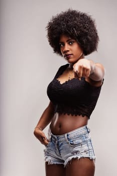 Young latin woman with afro hairstyle pointing her finger while posing confidently in a studio setting, wearing a black top and denim shorts, exuding a sense of empowerment and freedom