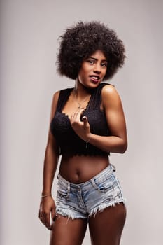 Studio portrait of a sensual young latin american woman with afro hair looking at camera, wearing a black top and denim shorts, posing with a diva attitude