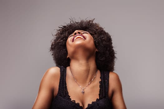 Studio portrait of a beautiful, smiling latin american woman with curly hair, wearing a black top and a necklace, laughing with her head tilted back, expressing joy and freedom