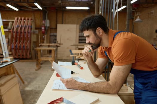 Pensive man carpenter reading blueprints analyzing and thinking about project during woodworking and furniture production process at workshop