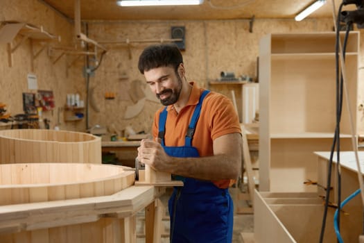 Carpenter working on mini home wooden phytobarrel sauna at workshop. Artisan enjoying woodworking process creating compact steam room for customer