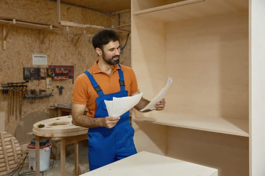 Carpenter looking at paper sheet with project blueprint reading assembly instructions standing over woodshop interior