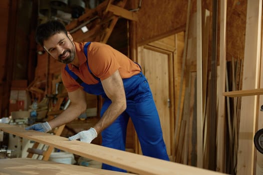 Young man carpenter in uniform putting wood plank on modern equipment closeup view. Experienced workman enjoying DIY woodworking project at his own small furniture factory