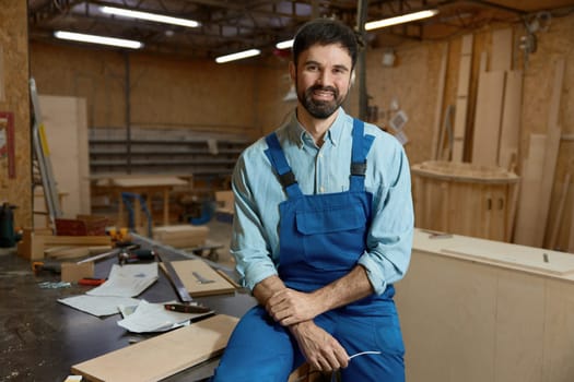 Skilled young adult woodworker sitting on workbench in well-equipped workshop studio. Man artisan smiling looking at camera