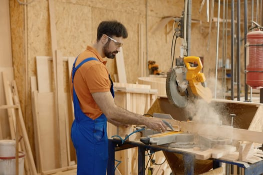 Skilled concentrated male carpenter wearing coveralls and protective glasses sawing wood plank operating with round circular saw at carpentry workbench