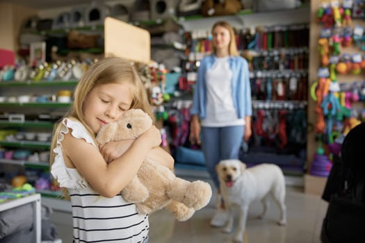 Little girl hugging soft bunny toy standing over pet store with mother and dog on blurred background