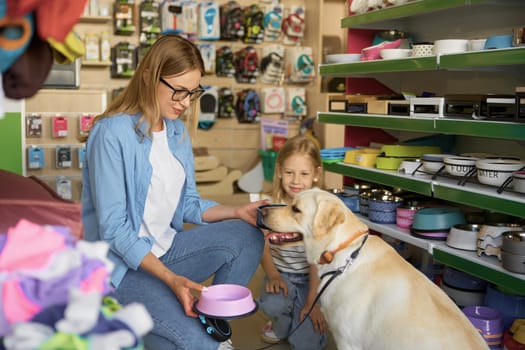 Mother and toddler daughter choosing new plastic food plate for Labrador dog enjoying shopping at pet store