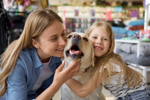Portrait of cheerful smiling mother and daughter cuddling with dog over pet shop background. Happy family shopping time at showroom for domestic animals