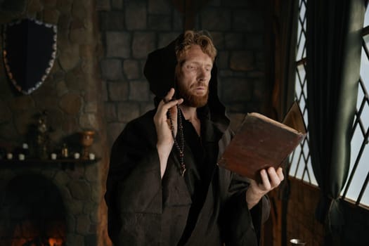 Medieval monk wearing robe with hood reading sacred religious book praying with rosary beads in hand