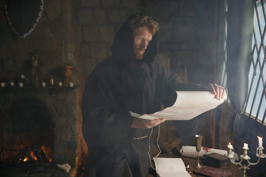 Mature medieval monk wearing robe with hood reading sacred manuscript on parchment holding historical religious document in hands while sitting at table in monastery cell