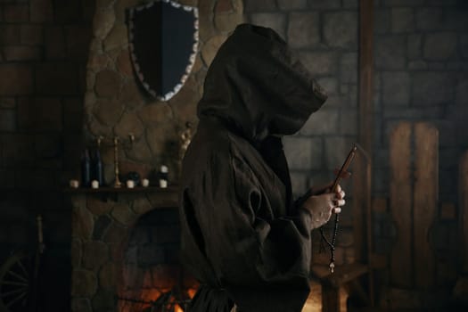 Faithful priest in robe hood praying with rosary beads and cross over ancient monastery interior. Side view portrait