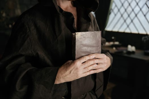 Mature priest wearing medieval robe clothing holding closed bible book and rosary beads in hands closeup