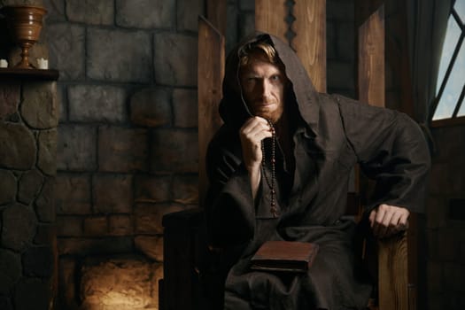 Portrait of medieval monk with mysterious look wearing robe hood sitting on wooden armchair over ancient monastery background