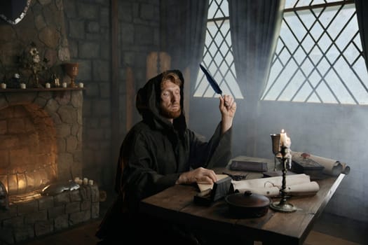 Medieval monk in cassock with quill pen writing historic manuscripts at table under candlelight in abbey cell