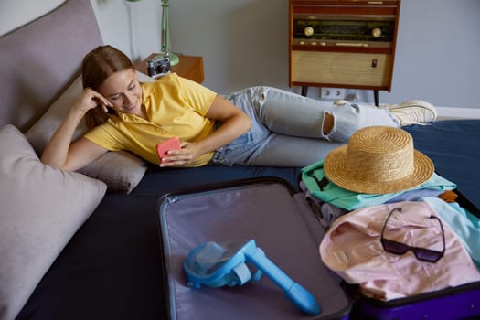 Young woman lying on bed with opened travel suitcase. Pretty girl wishing about luxurious days at tropical resort