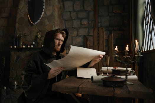 Medieval monk wearing robe with hood reading sacred manuscript on parchment holding historical religious document in hands while sitting at table in monastery cell
