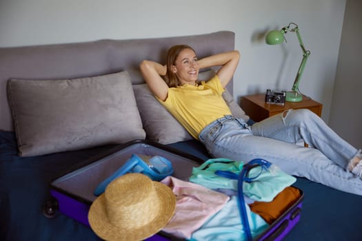 Dream-like young woman lying on bed with opened travel suitcase. Pretty girl wishing about luxurious days at tropical resort
