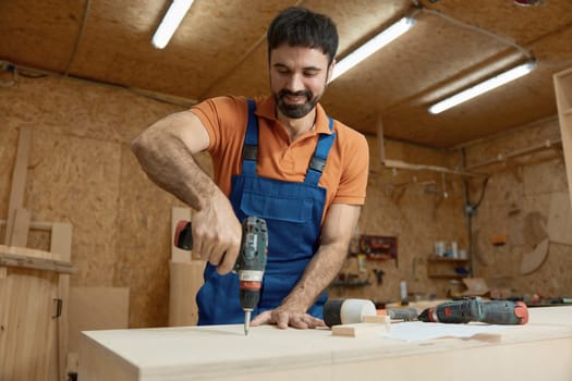 Male carpenter working with drill electric screwdriver making holes in wooden plank. Woodworking and furniture production process