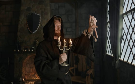 Portrait of hooded monk in medieval costume holding wooden cross and candlestick praying in church