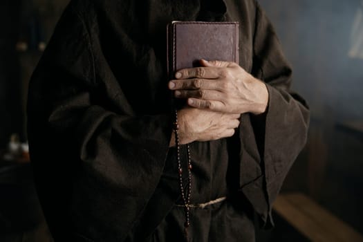Priest wearing medieval robe clothing holding closed bible book and rosary beads in hands closeup