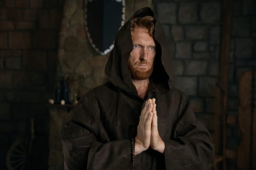 Portrait of blessed priest praying having faith believe in God. Monk wearing robe with hood holding folded hands front of chest standing over quiet dark mystical monastic background