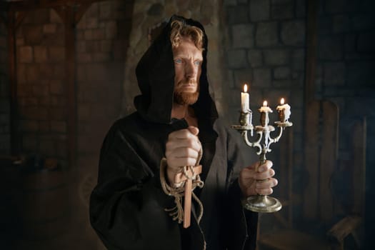 Portrait of hooded monk in medieval costume holding wooden cross and candlestick praying in church