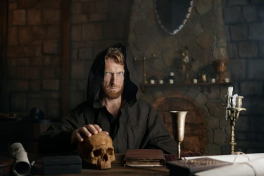 Portrait of mystical spooky medieval monk in black cassock putting hands on human skull prefroming spiritualistic seance or ritual. Fetal stage