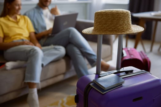 Closeup view straw hat, passport and sunglasses on travel baggage packed for vacation with happy married couple sitting and browsing internet on laptop