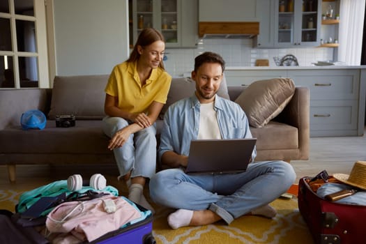 Loving couple booking vacation trip choosing flight on internet via laptop while sitting in home living-room with opened travel suitcases
