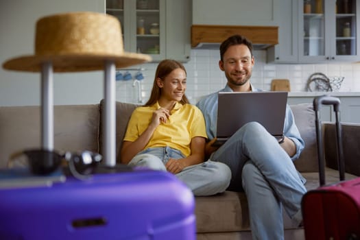 Happy loving couple planning honeymoon tour searching flight ticket or hotel for reservation on internet while sitting in home living-room with packed travel suitcases