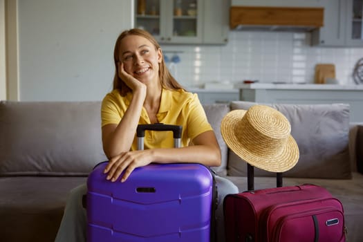 Happy woman wearing straw summer hat with travel luggage getting ready for vacation. Pretty girl tourist sitting on home sofa waiting for plane to tropical resort